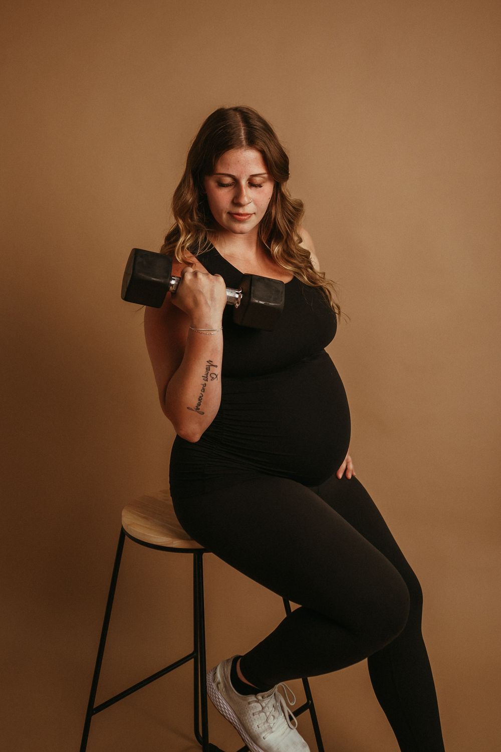 An expectant woman in black athletic wear holds a dumbbell while sitting on a stool against a warm brown backdrop.