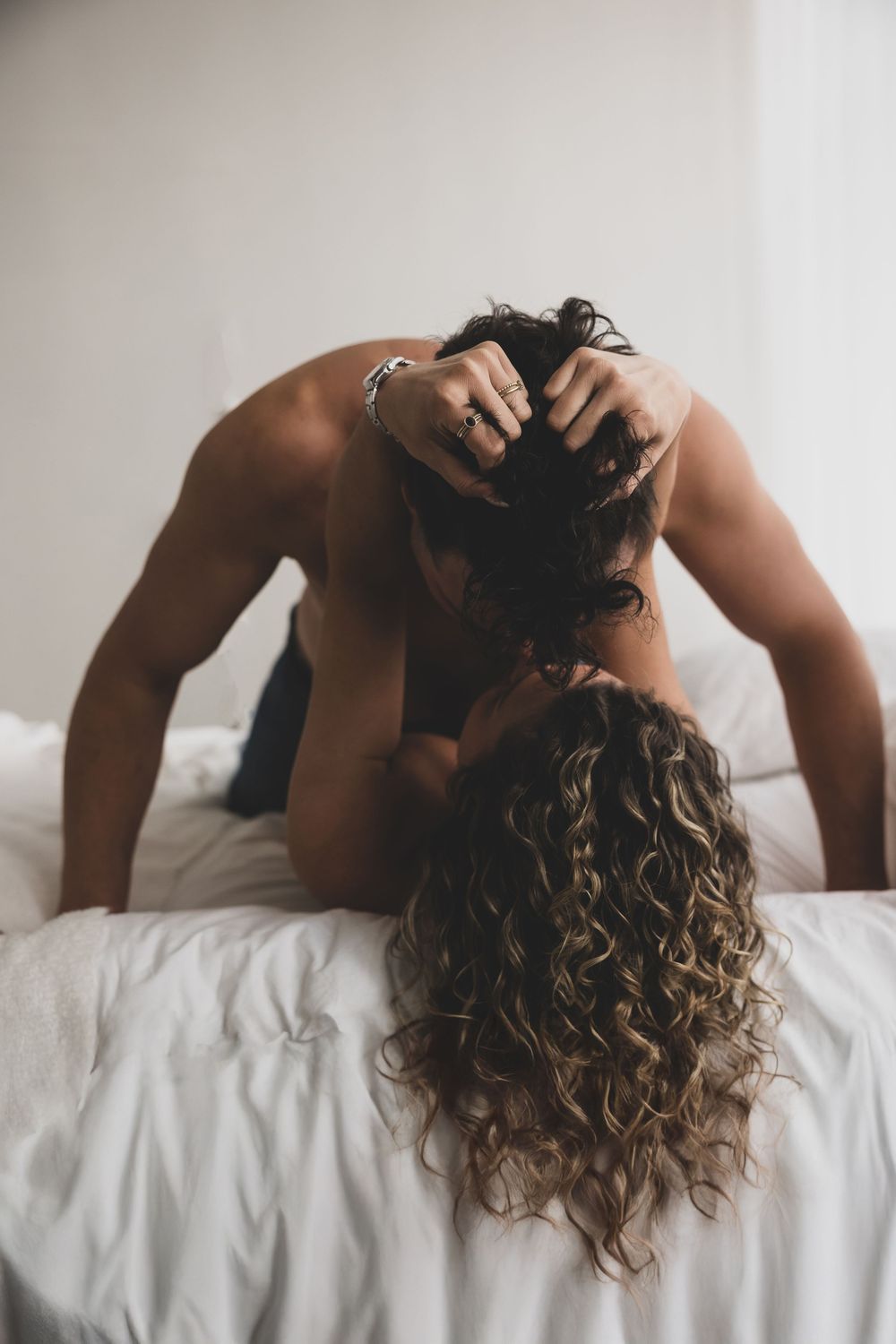 Person with curly highlighted hair lying back on white bedding in a relaxing boudoir style photoshoot series.