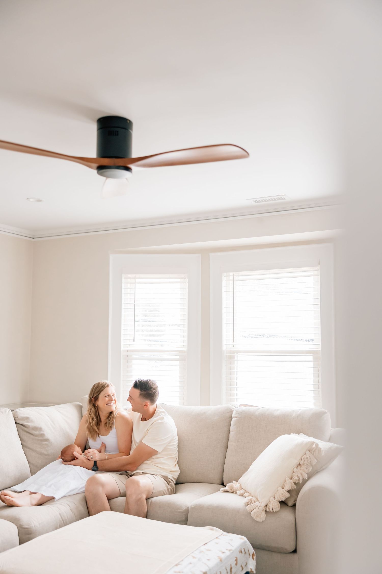 A modern living room with a sleek ceiling fan features a plush cream sectional sofa beneath bright windows.