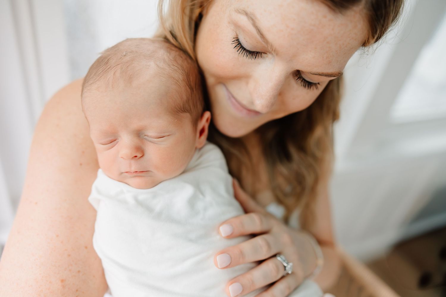 A tender moment between mother and sleeping newborn baby in white against soft light background.