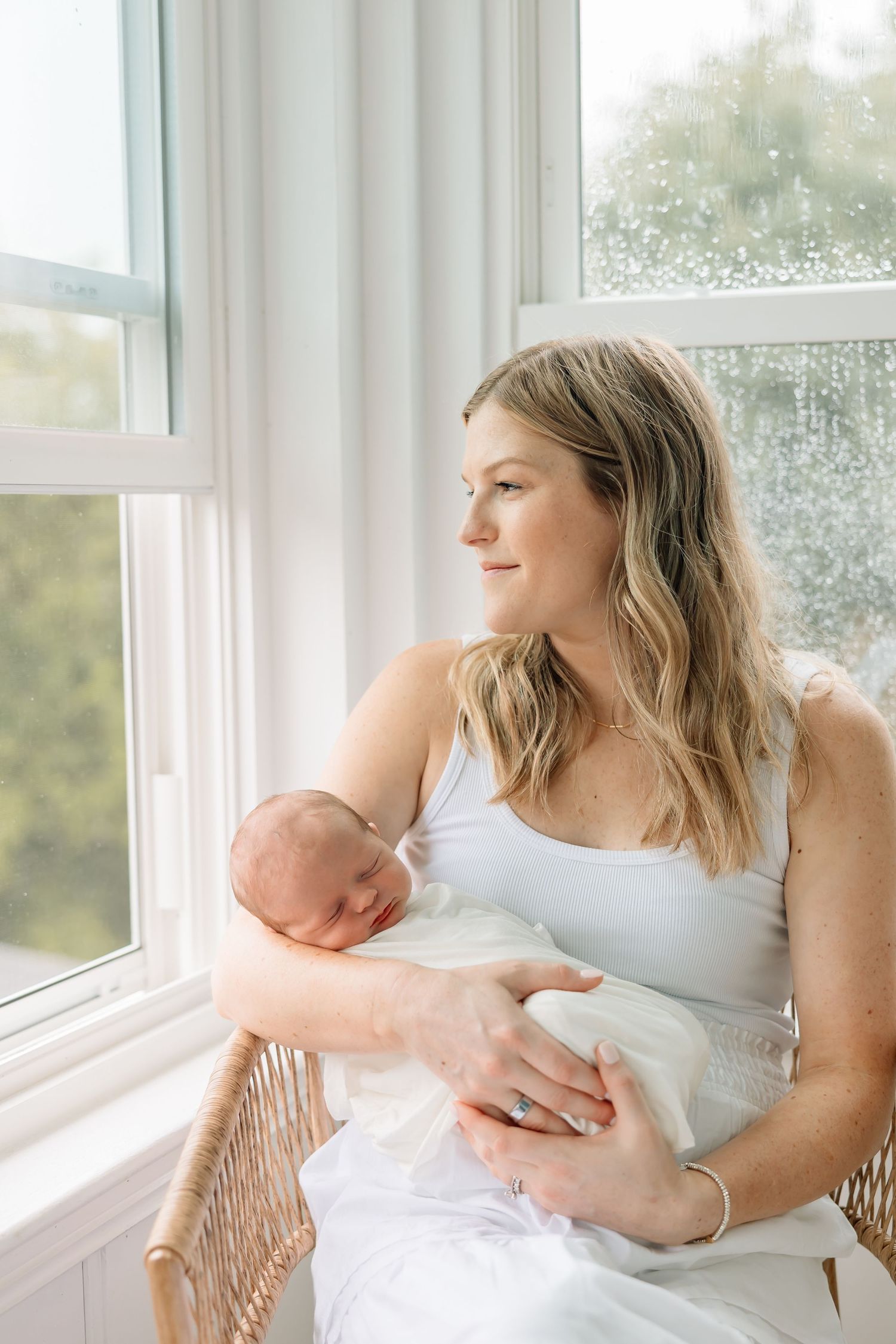 A mother in white sits by a window cradling her sleeping newborn baby in natural light.