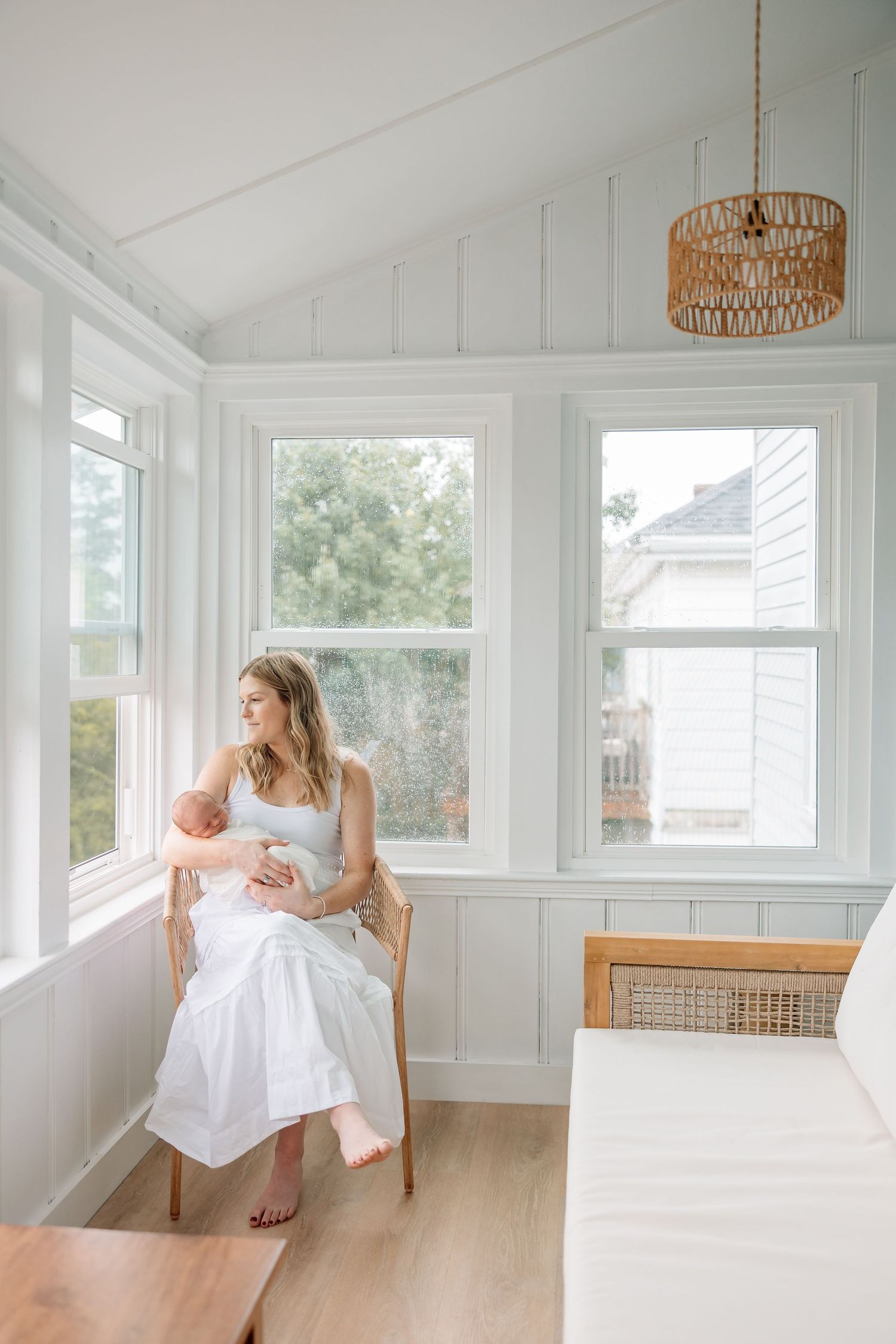 A bright sunlit room with white walls features rattan pendant lights and large windows overlooking greenery.