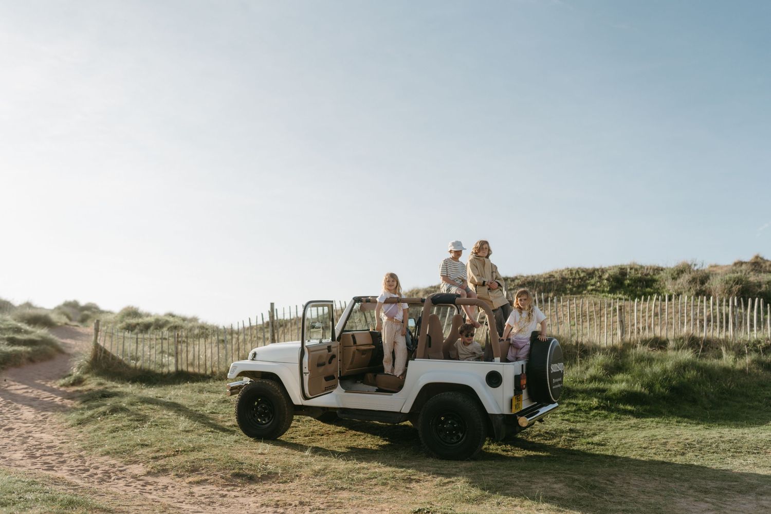 A white safari vehicle parked on a grassy coastal path with people and wooden fence rails in the background.