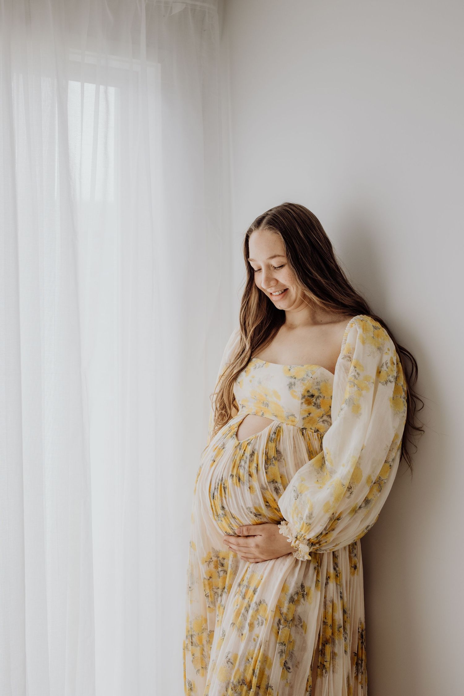 Expecting mother in floral yellow dress stands by window with hands cradling baby bump in soft natural light.