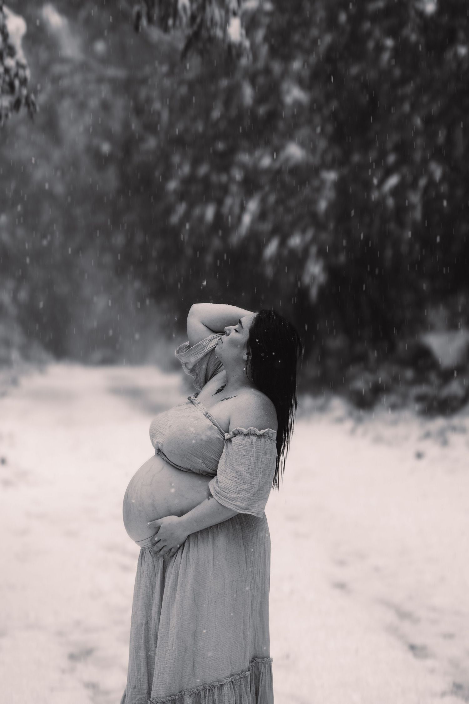 Black and white photo of a pregnant woman in a flowy dress standing in a snowy scene while holding her baby bump.