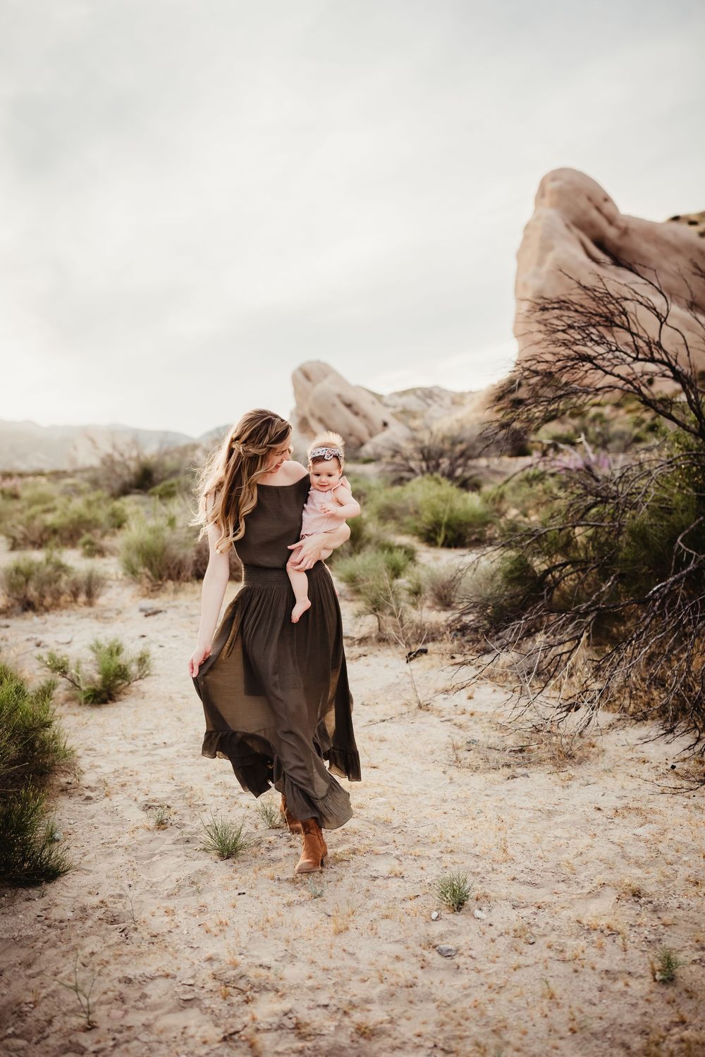 Family portrait in a desert landscape with rocky formations as woman in flowing dress holds baby against muted sky.