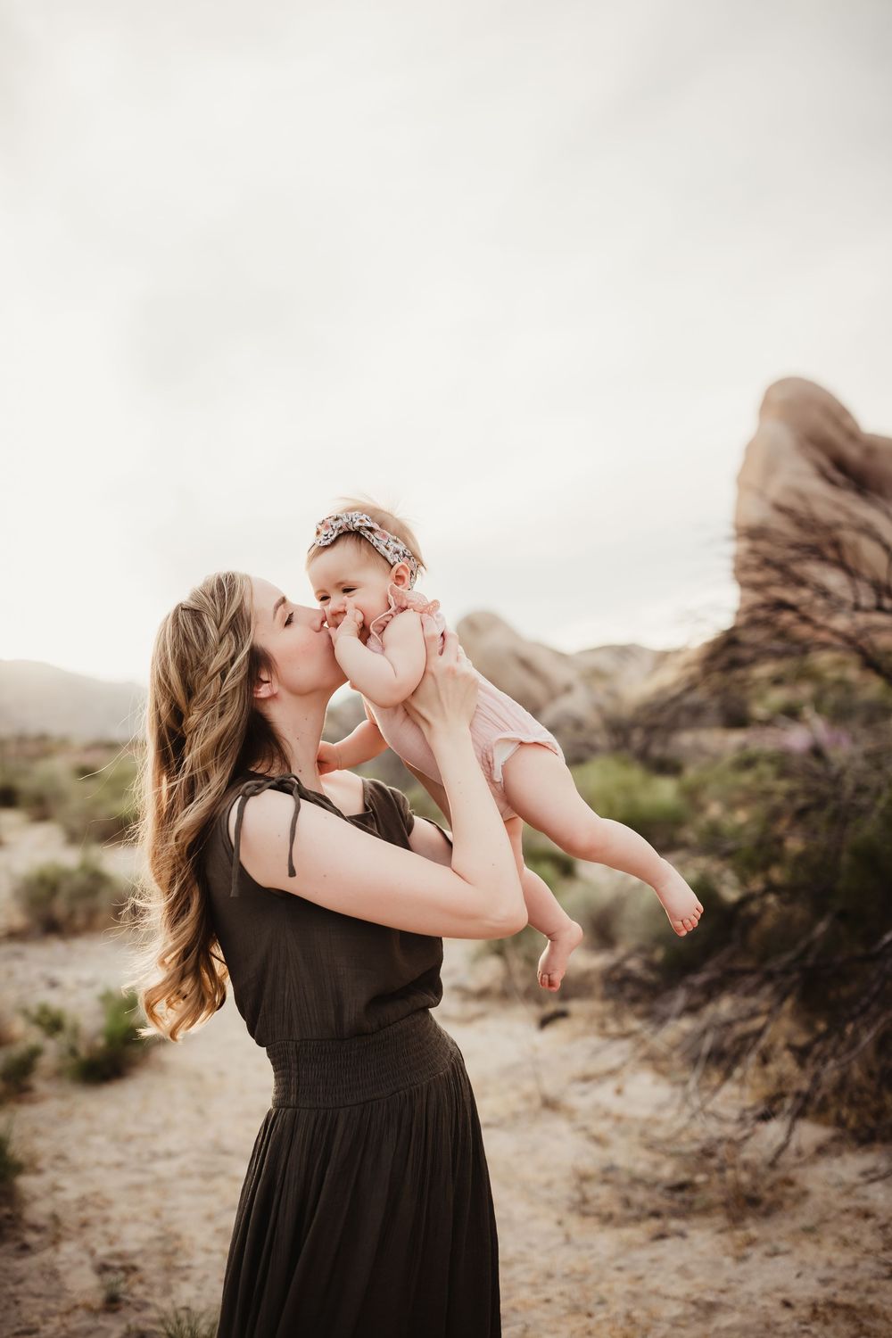 Tender moment between mother and baby amidst desert rock formations during sunset photoshoot.