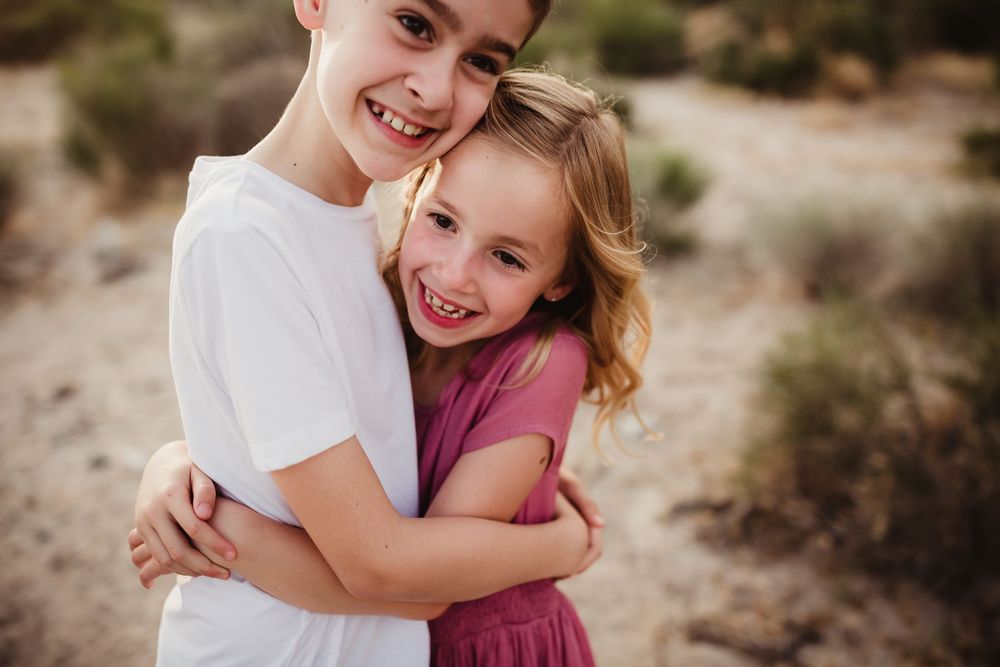 Two siblings share a joyful embrace while laughing in a desert setting.