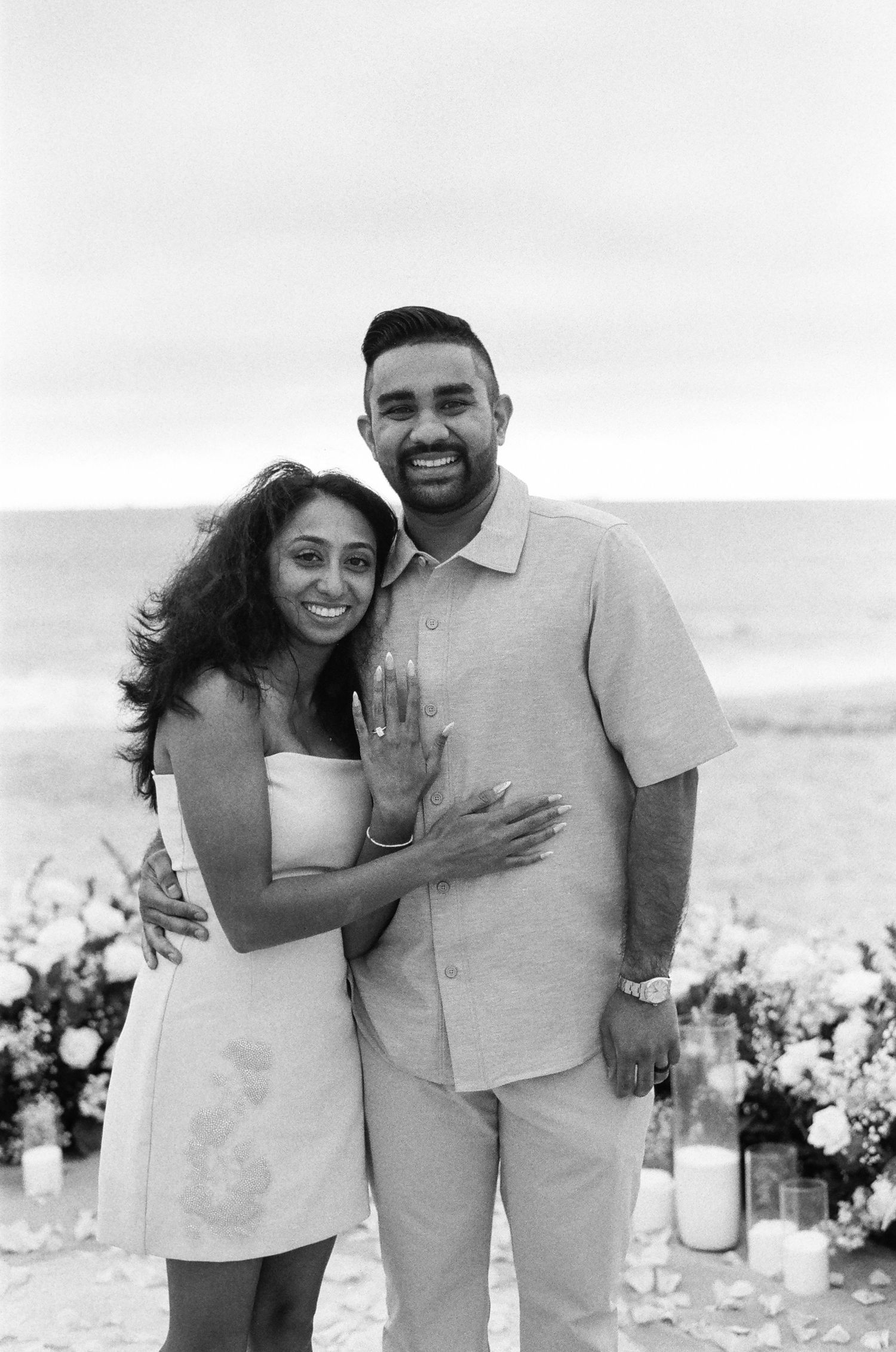 A happy couple embraces on the beach surrounded by flowers during what appears to be an engagement or wedding moment.