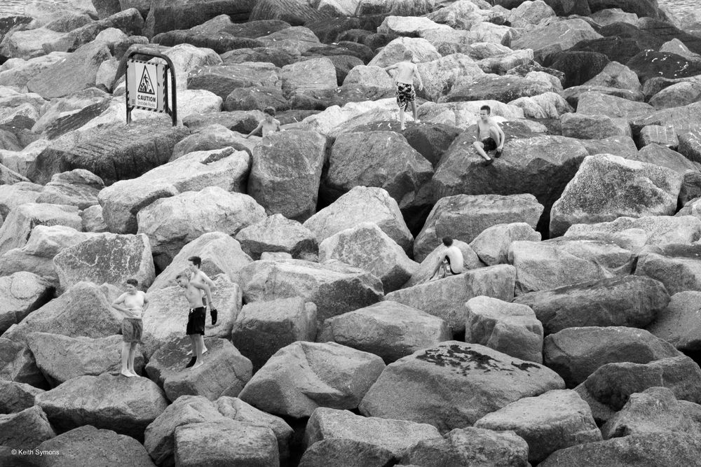 A black and white image of large jagged rocks forming a breakwater jetty along a shoreline.