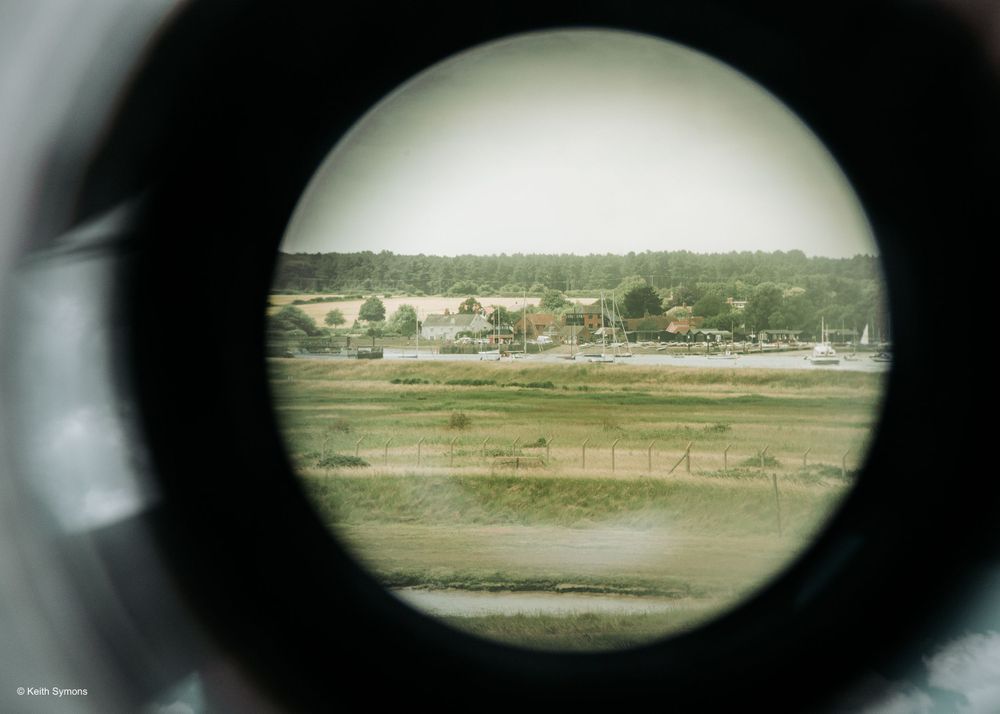 A rural landscape viewed through a circular telescope or spotting scope shows fields and buildings in the distance.