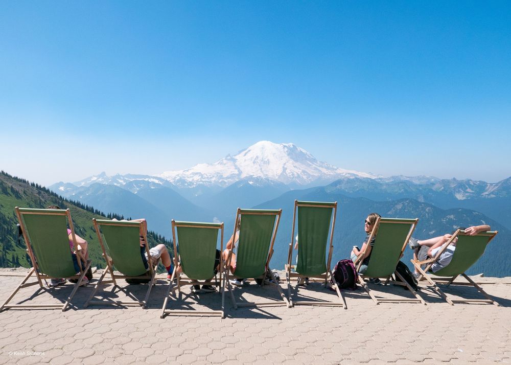 People relax in green lounge chairs overlooking Mount Rainier and a scenic mountain landscape on a clear summer day.