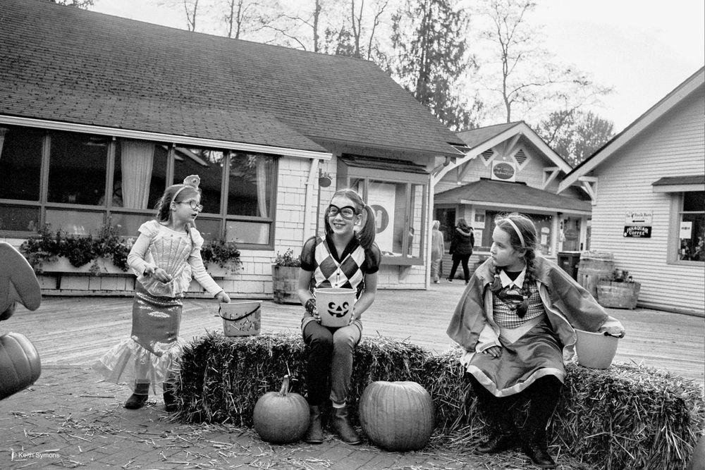 A black and white photo of three children sitting on hay bales with pumpkins in front of a house.