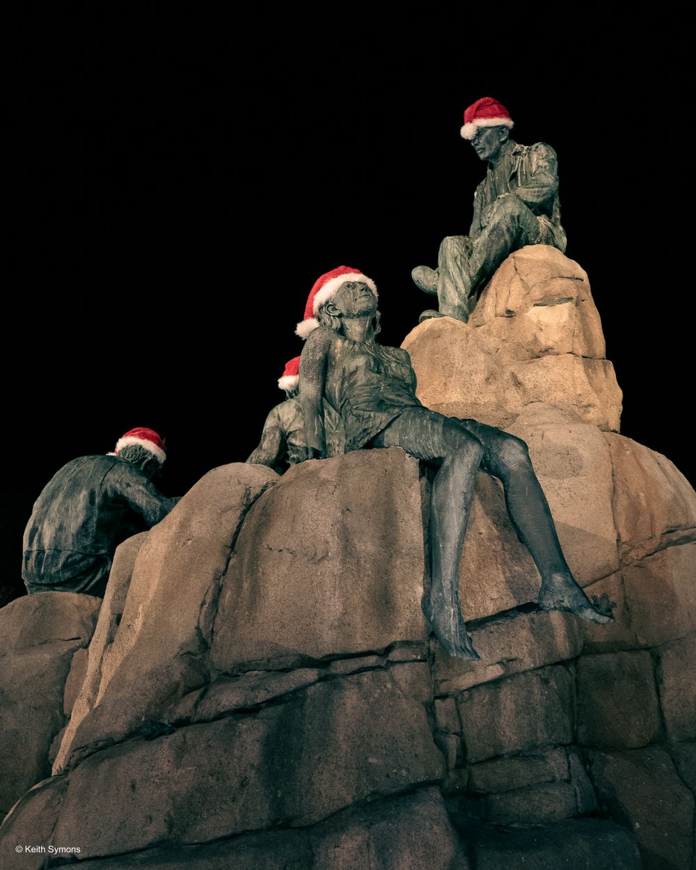 Three rock climbers wearing Santa hats perch on a rocky formation against a dark background.