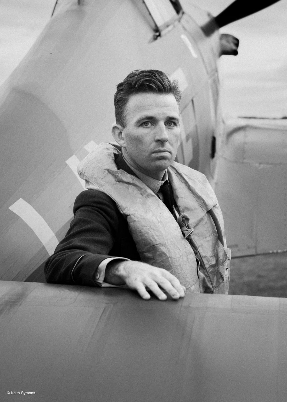 A black and white portrait of an aviator in flight gear standing beside an aircraft.