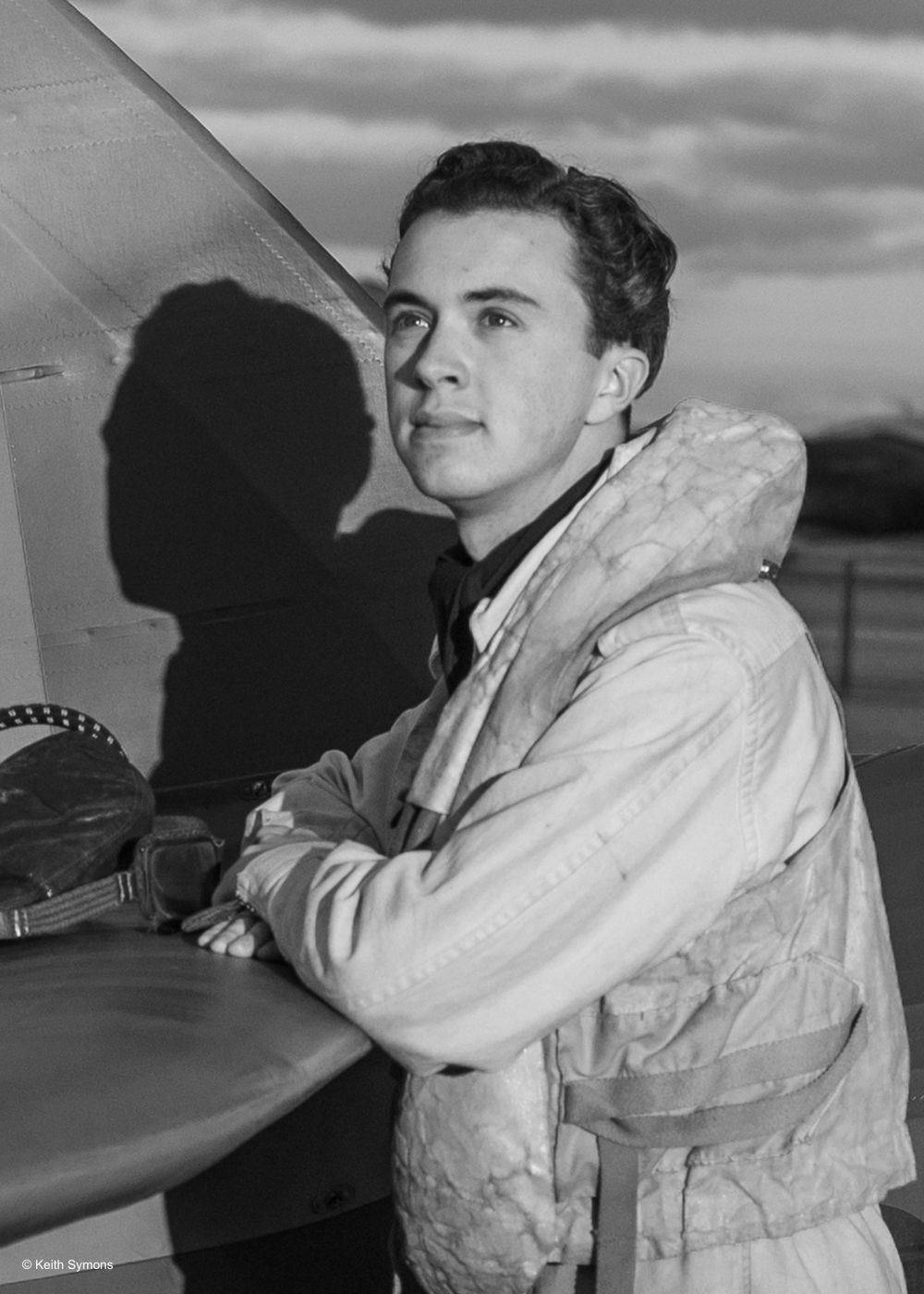 A black and white photograph of an aviator wearing a light-colored flight jacket standing next to an aircraft.