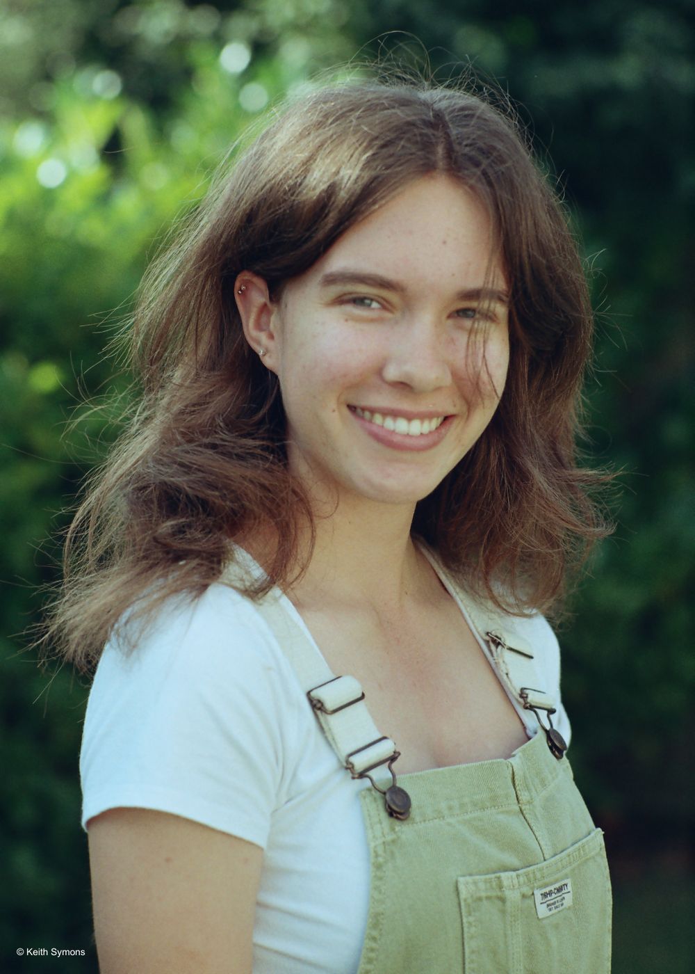 Portrait of someone in a white shirt and overalls smiling outdoors with green foliage in background.