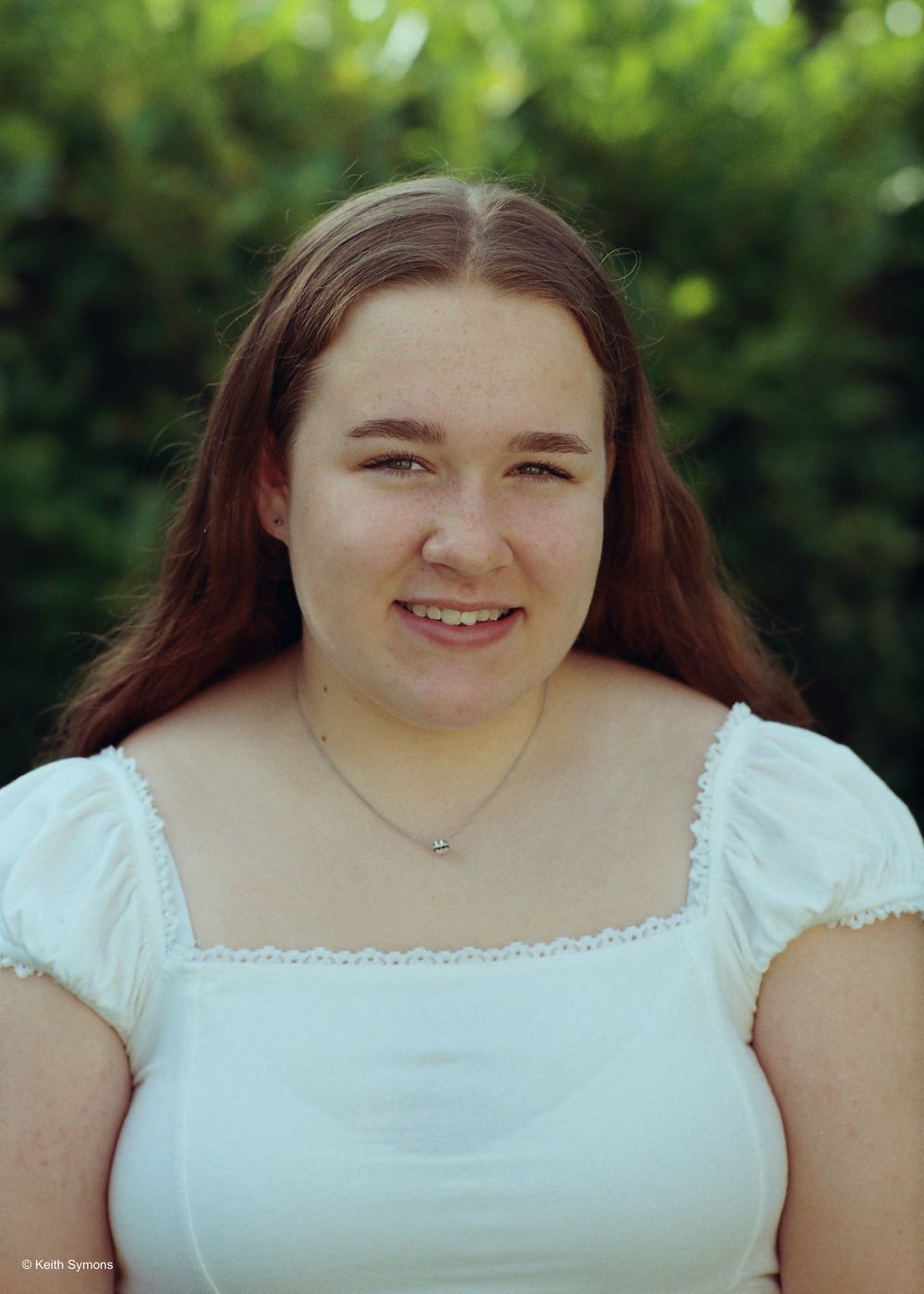 An outdoor portrait showing someone in a white square-neck top with short sleeves against a blurred green background.