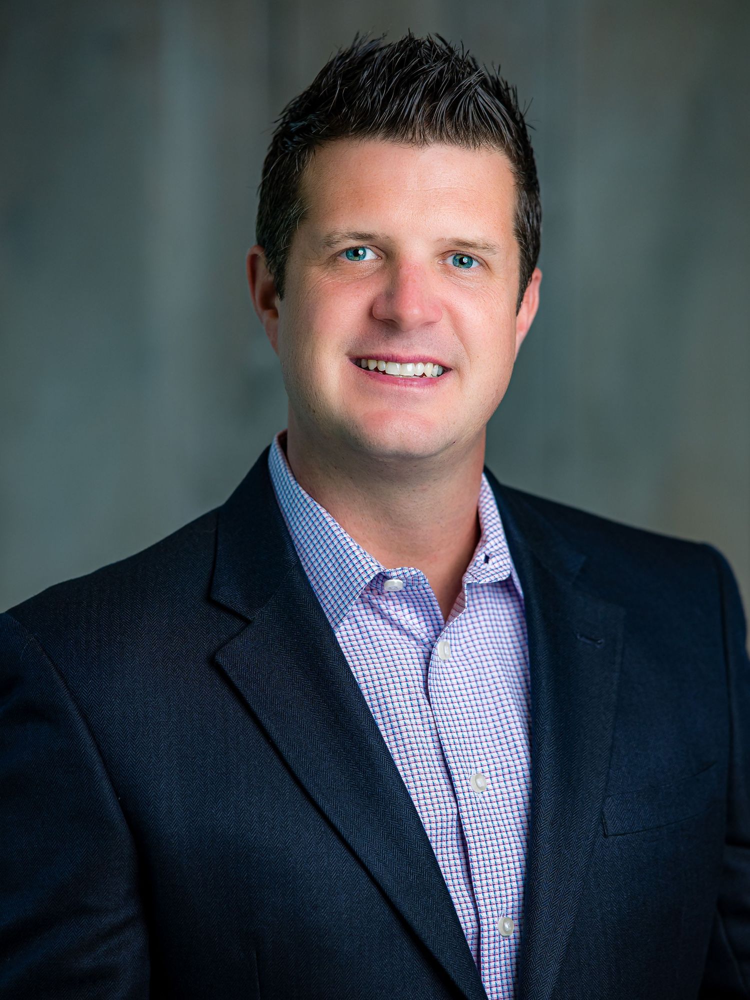 Professional headshot of person in navy blazer and purple checkered shirt against blurred background.