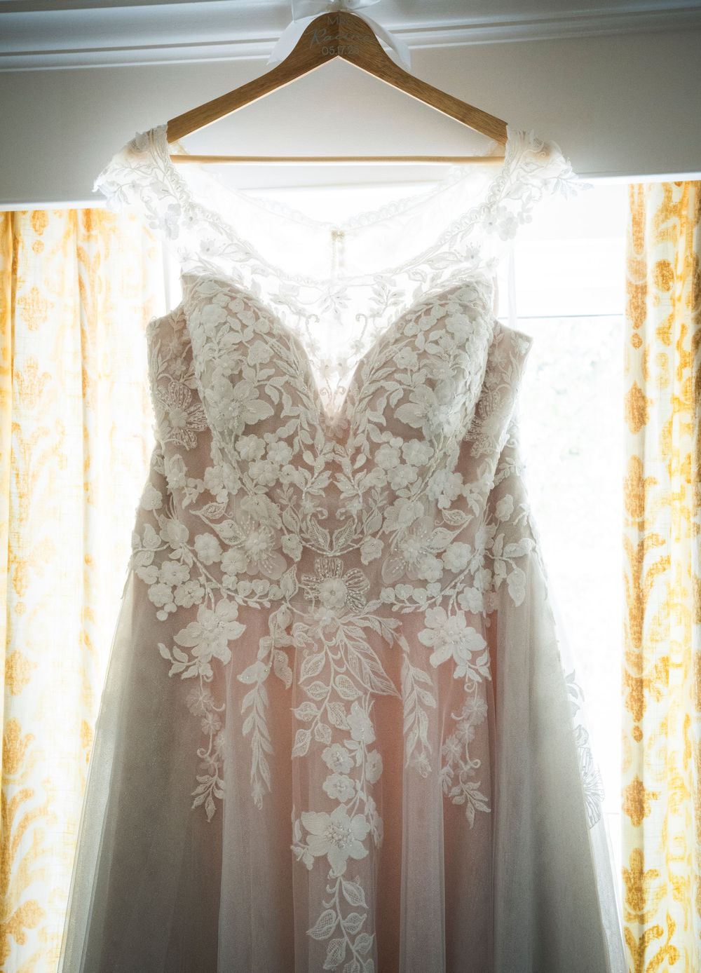 A delicate white lace wedding dress with floral appliques hangs on a wooden hanger against sheer curtains in natural light.