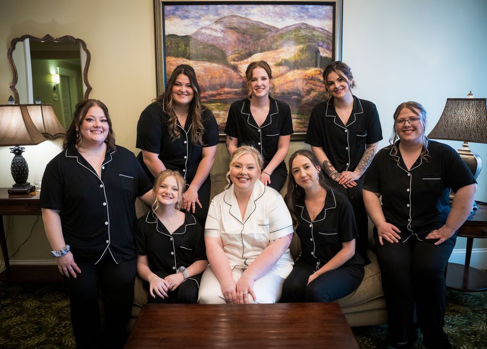 A wedding party in matching black and white attire poses together in front of a landscape painting.