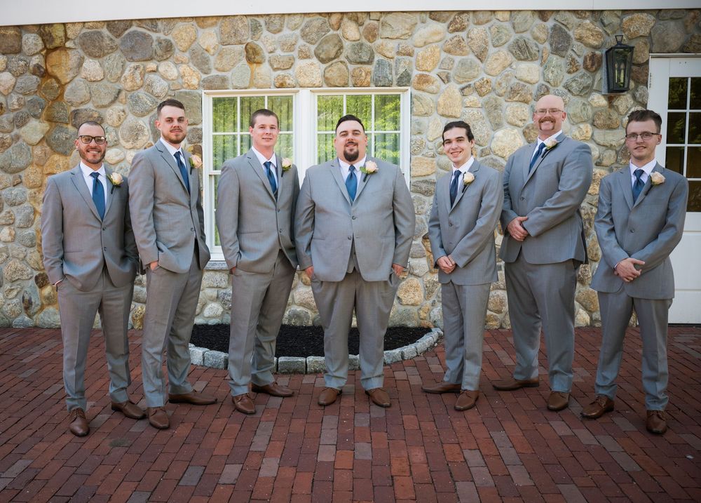A lineup of groomsmen wearing matching light gray suits standing together in front of a stone wall during a wedding.