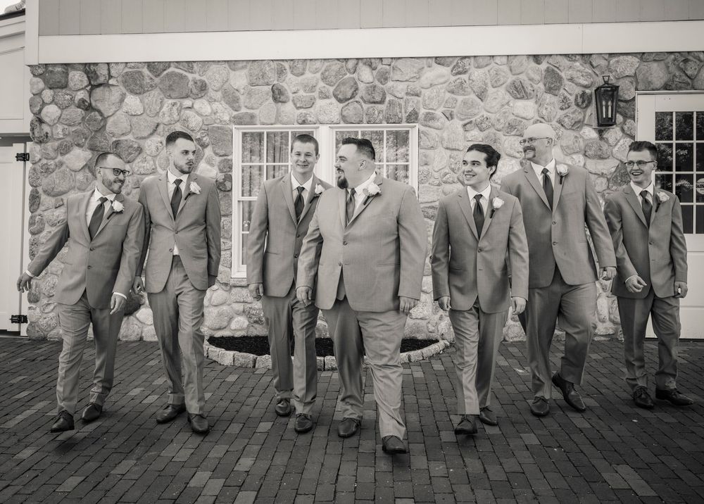 A group of groomsmen in matching gray suits pose together outside a stone building wall.