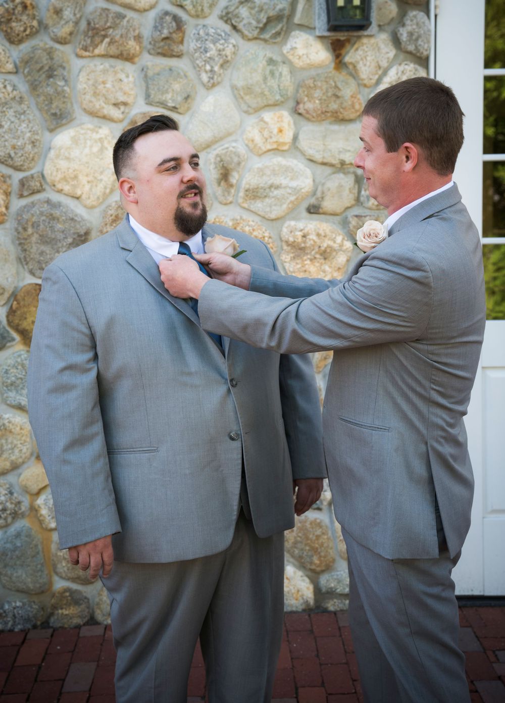 Two people in grey suits help each other adjust boutonnieres in front of a stone wall.