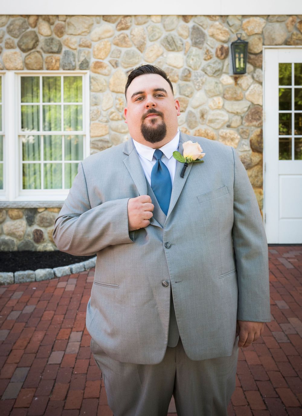 Groom in light grey suit with blue tie and boutonniere poses in front of stone wall with white windows.
