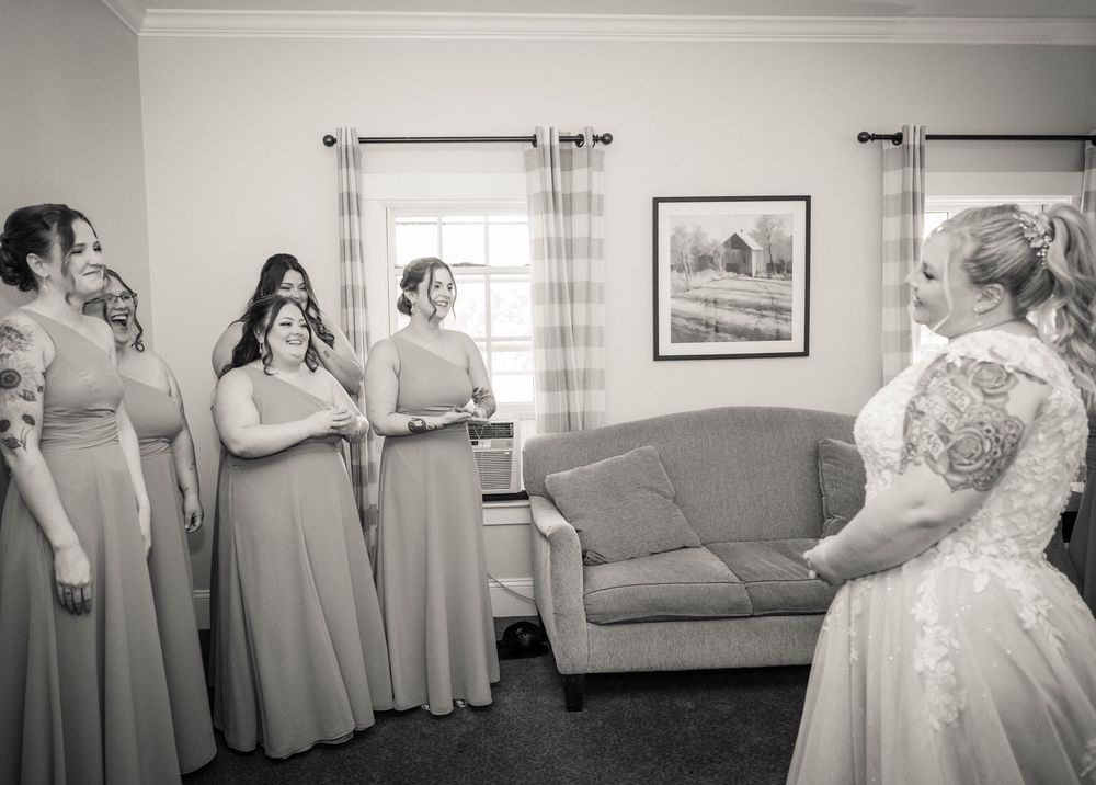 Black and white photo of bridesmaids in matching dusty rose gowns reacting emotionally to bride in lace wedding dress.