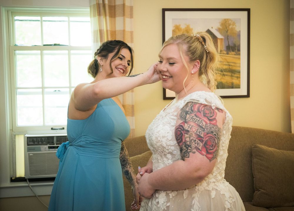 Two people share a joyful moment as one helps adjust the other's wedding dress in a sun-lit room.