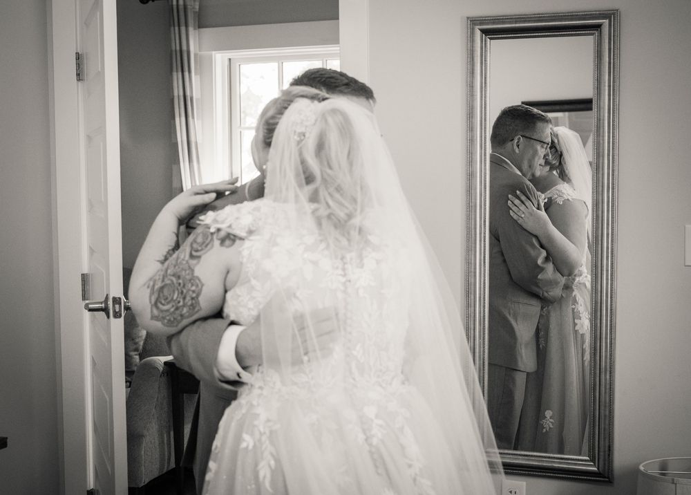 A bride in a white gown with tattoos looks in a mirror while getting ready on her wedding day.