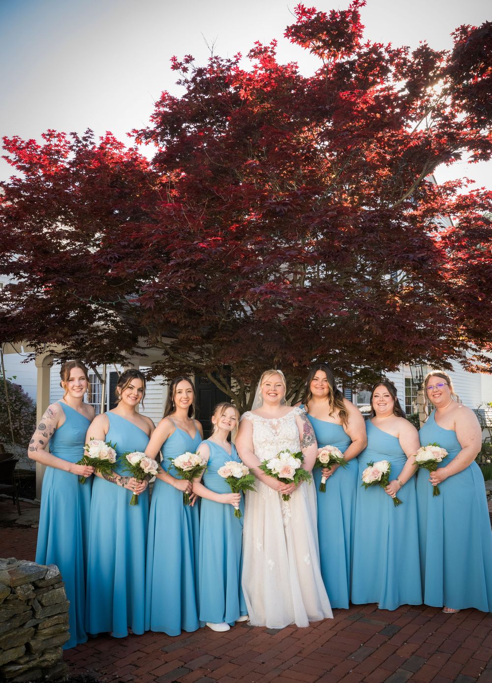 Wedding party posing beneath a vibrant red maple tree while holding bouquets in matching blue bridesmaid dresses.