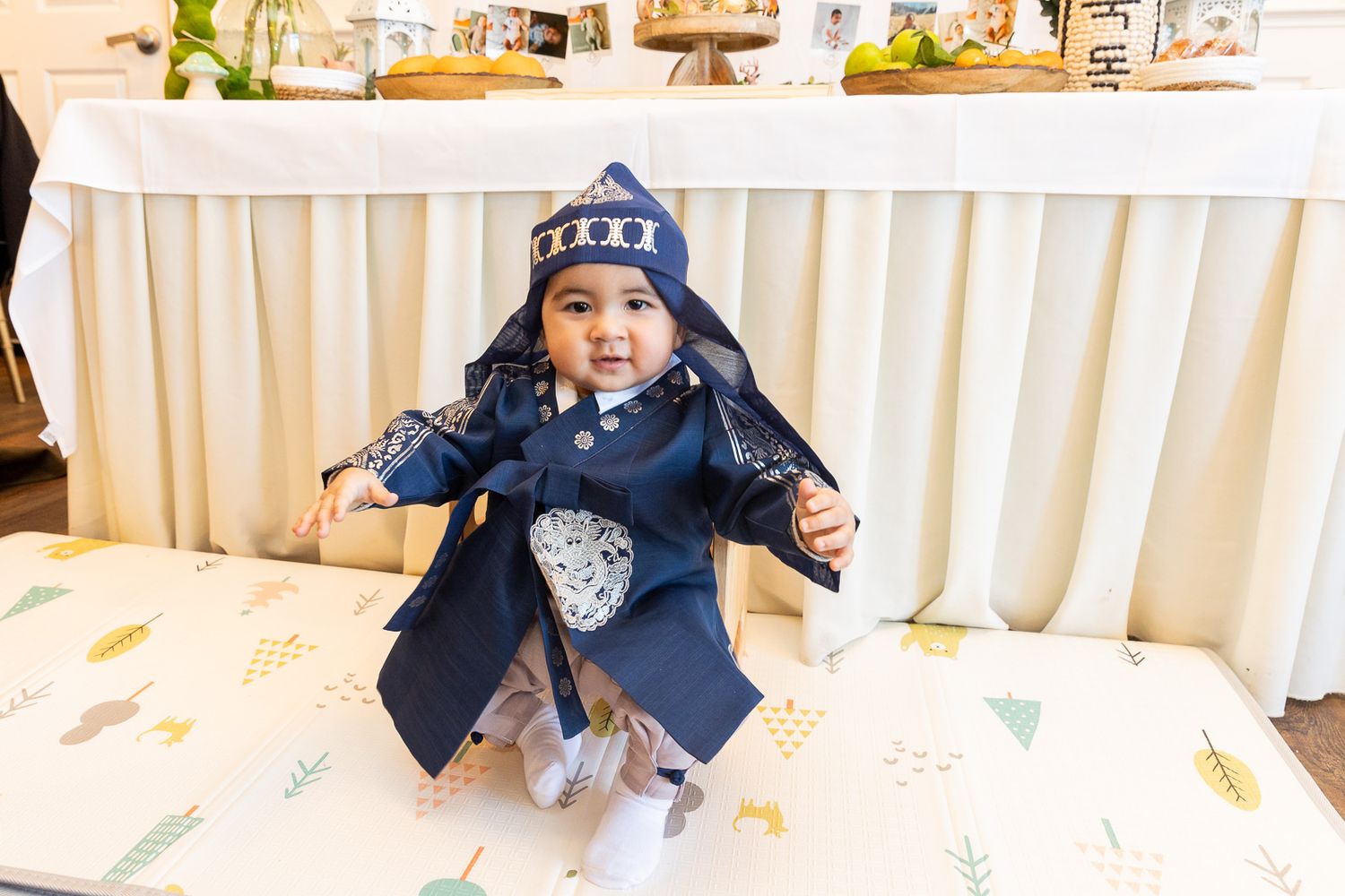 A young child wearing a navy blue coat and winter hat sits on a white couch at what appears to be a special event.