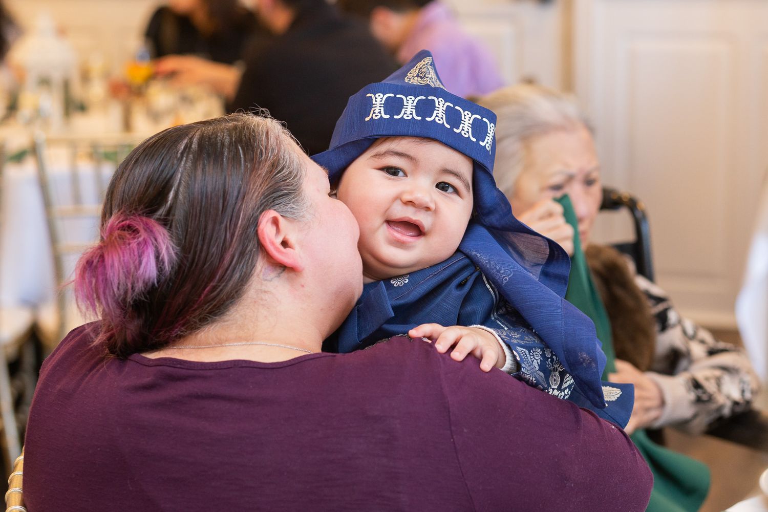 A joyful baby wearing a blue star-patterned hat smiles while being held at a gathering.