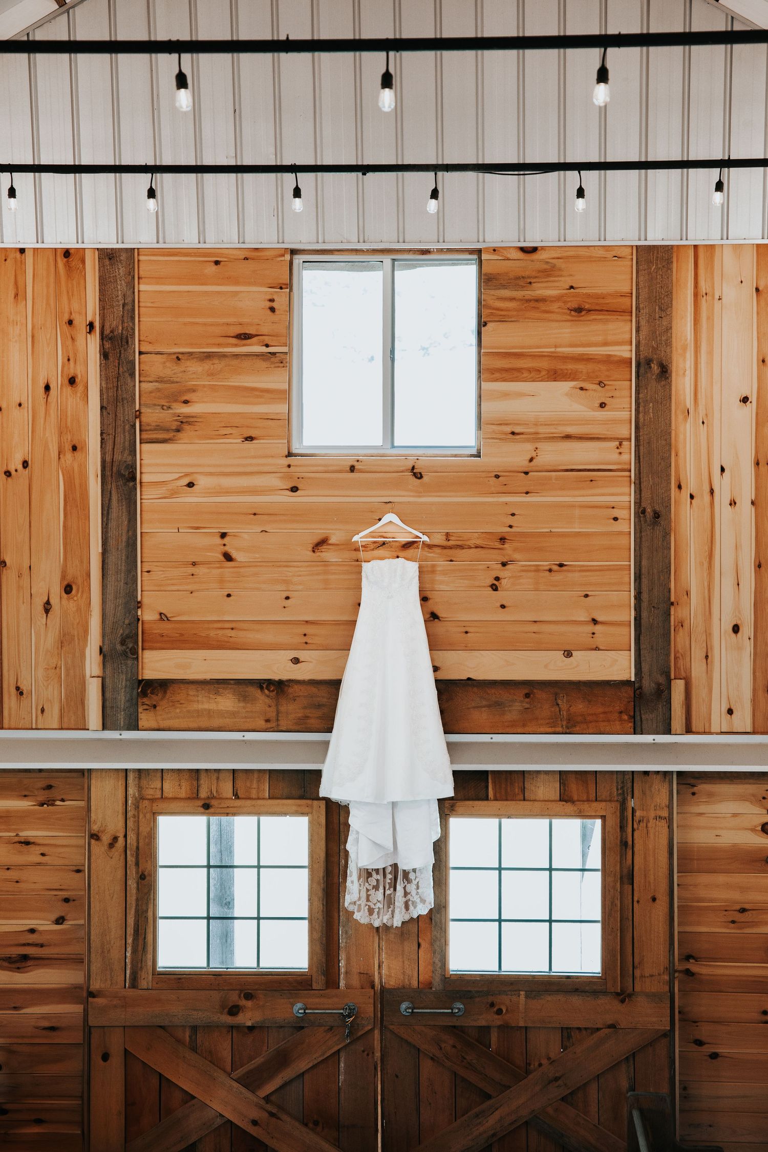 A white wedding dress hangs from a wooden hanger against rustic barn wood walls with windows and overhead string lights.