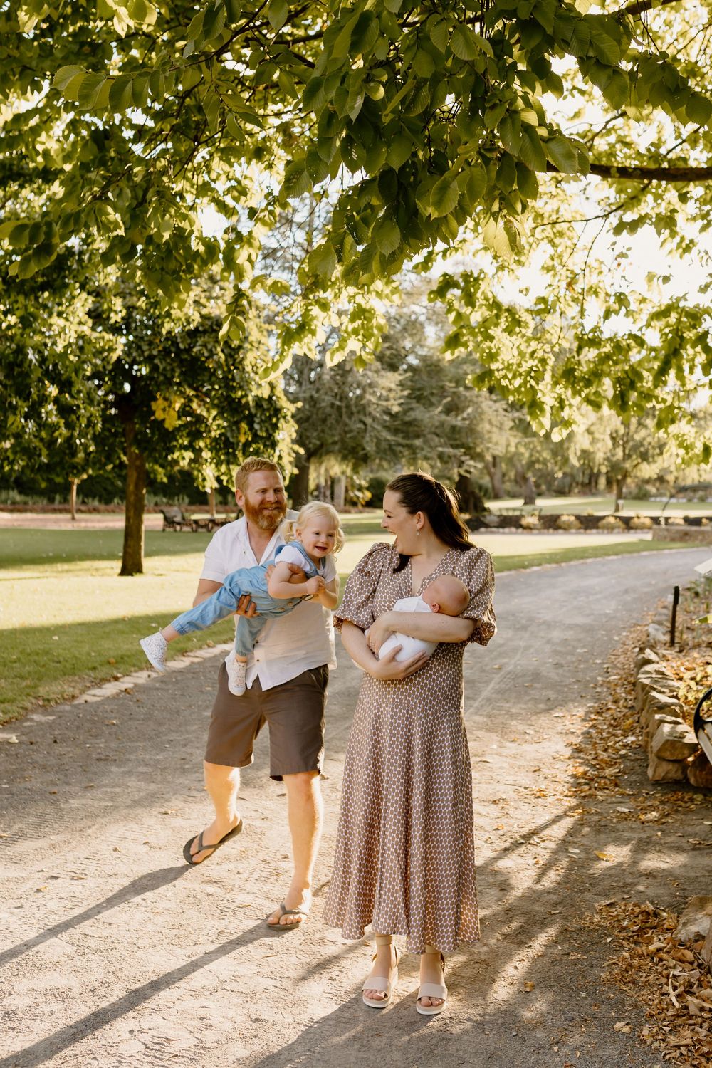 A couple walking along a sunlit path in a park holding their babies on a warm afternoon.