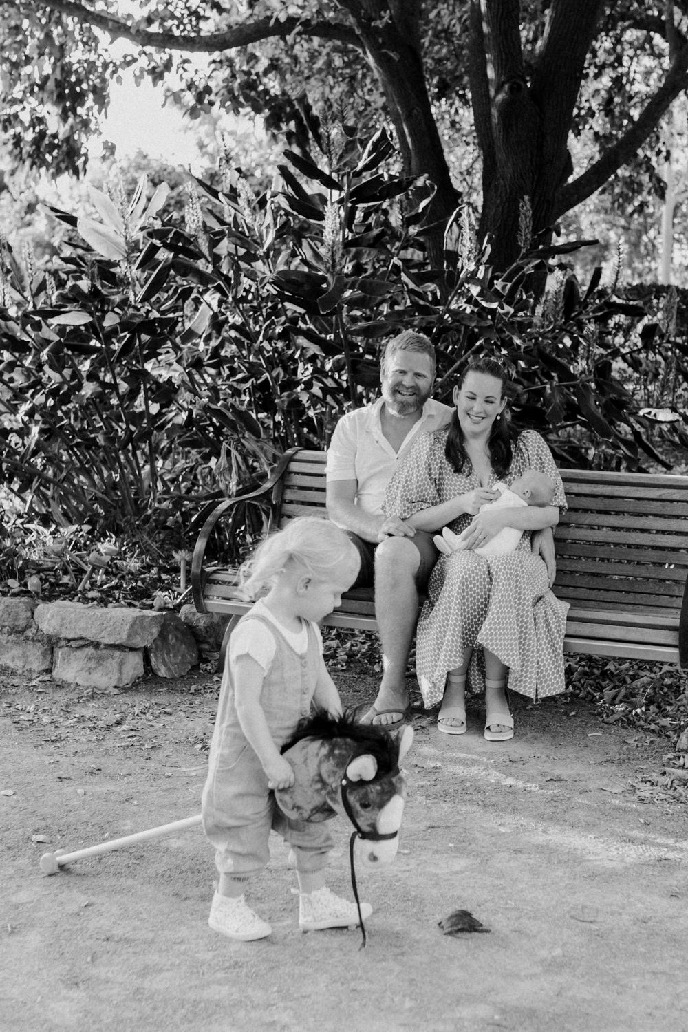 Black and white photo of people sitting on a garden bench while a small child plays with a dog on the ground.