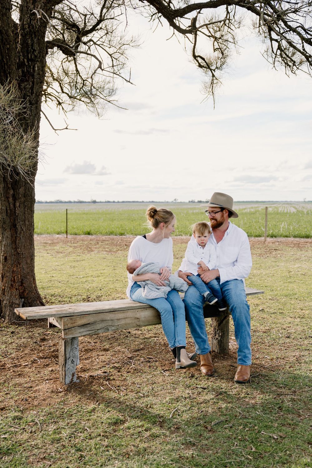 A family sits together on a rustic wooden bench under a large tree in an open rural field on a cloudy day.