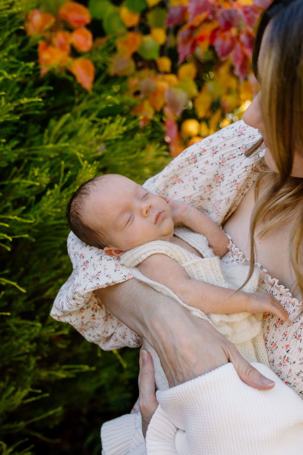 A sleeping baby in white lace clothing rests against someone wearing a matching lace dress outdoors with autumn foliage.