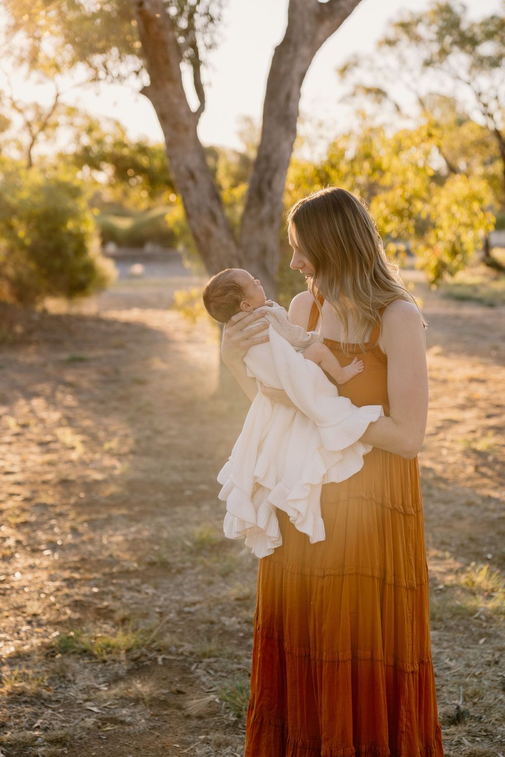 A woman in a flowing maxi dress holds a newborn baby while standing on a sunlit dirt path among trees.