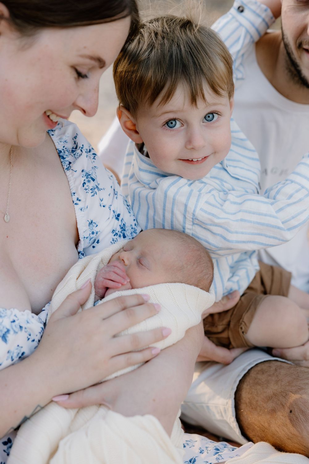 Family bonding moment as mother in floral dress cradles newborn baby while toddler sibling watches with bright eyes.