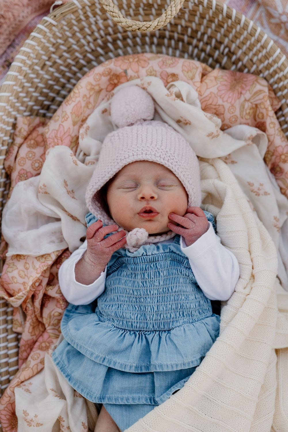 Newborn baby wearing a pink knit hat and denim outfit laying on a floral blanket.