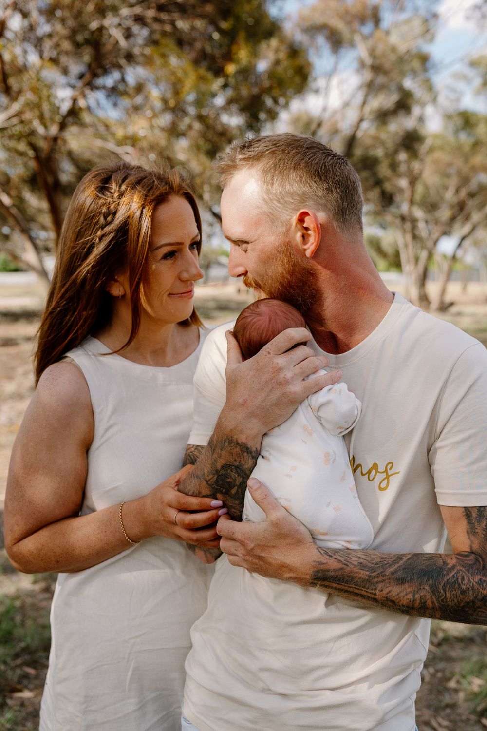 Loving couple in white outfits gazes adoringly at their newborn baby in an outdoor rural setting.