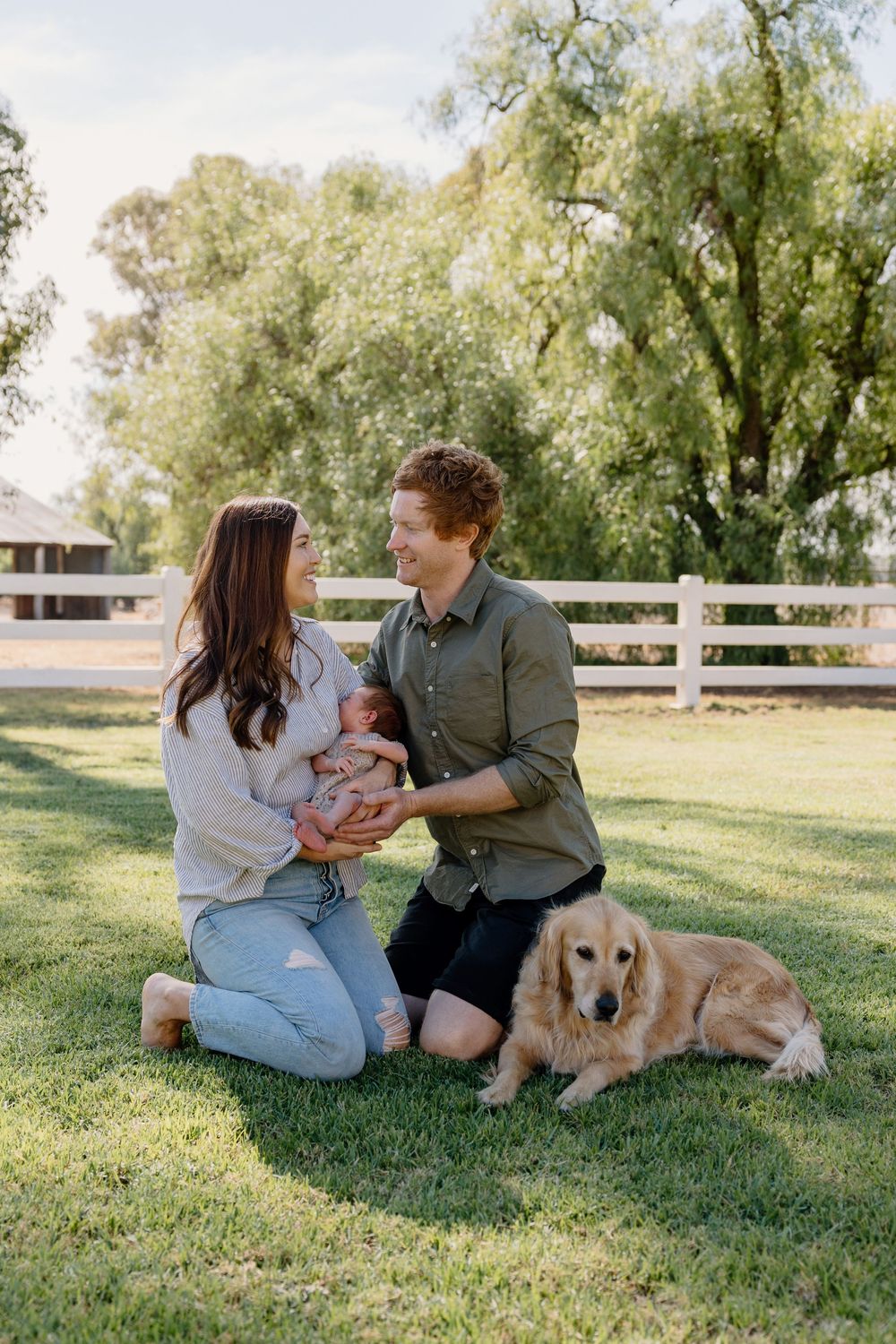 A couple with baby and golden retriever relaxing outdoors on grassy lawn by white fence on sunny afternoon.