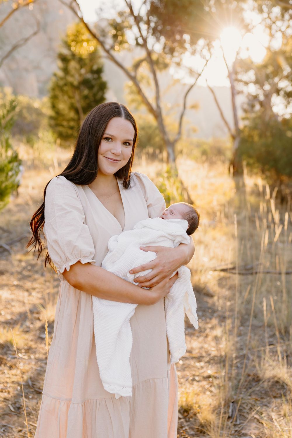 A person in a light flowy dress holds a swaddled newborn baby outdoors during golden hour sunset.