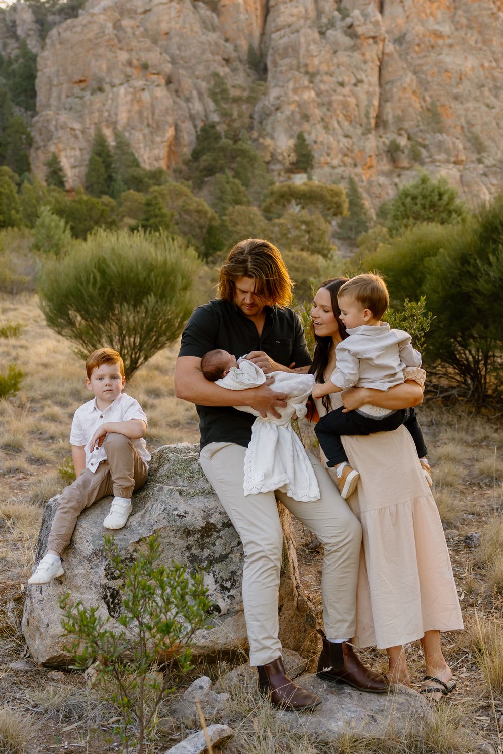 Family gathering outdoors at sunset against dramatic rocky cliffs with desert brush in natural landscape setting.