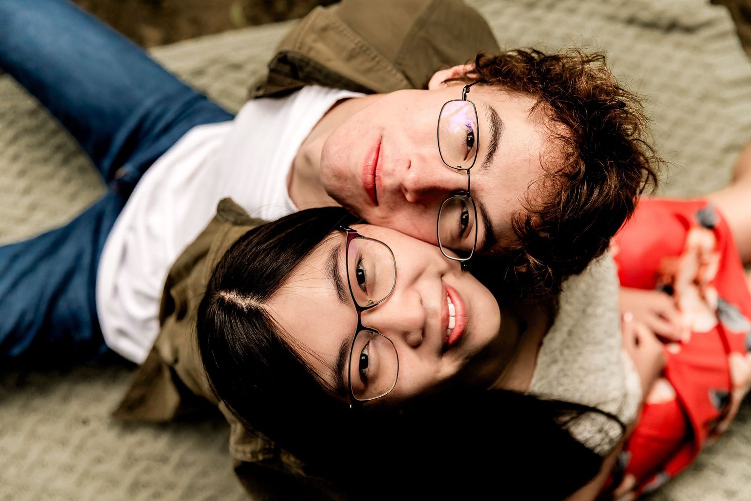 Two people wearing glasses lay on a grey blanket while smiling and relaxing together.
