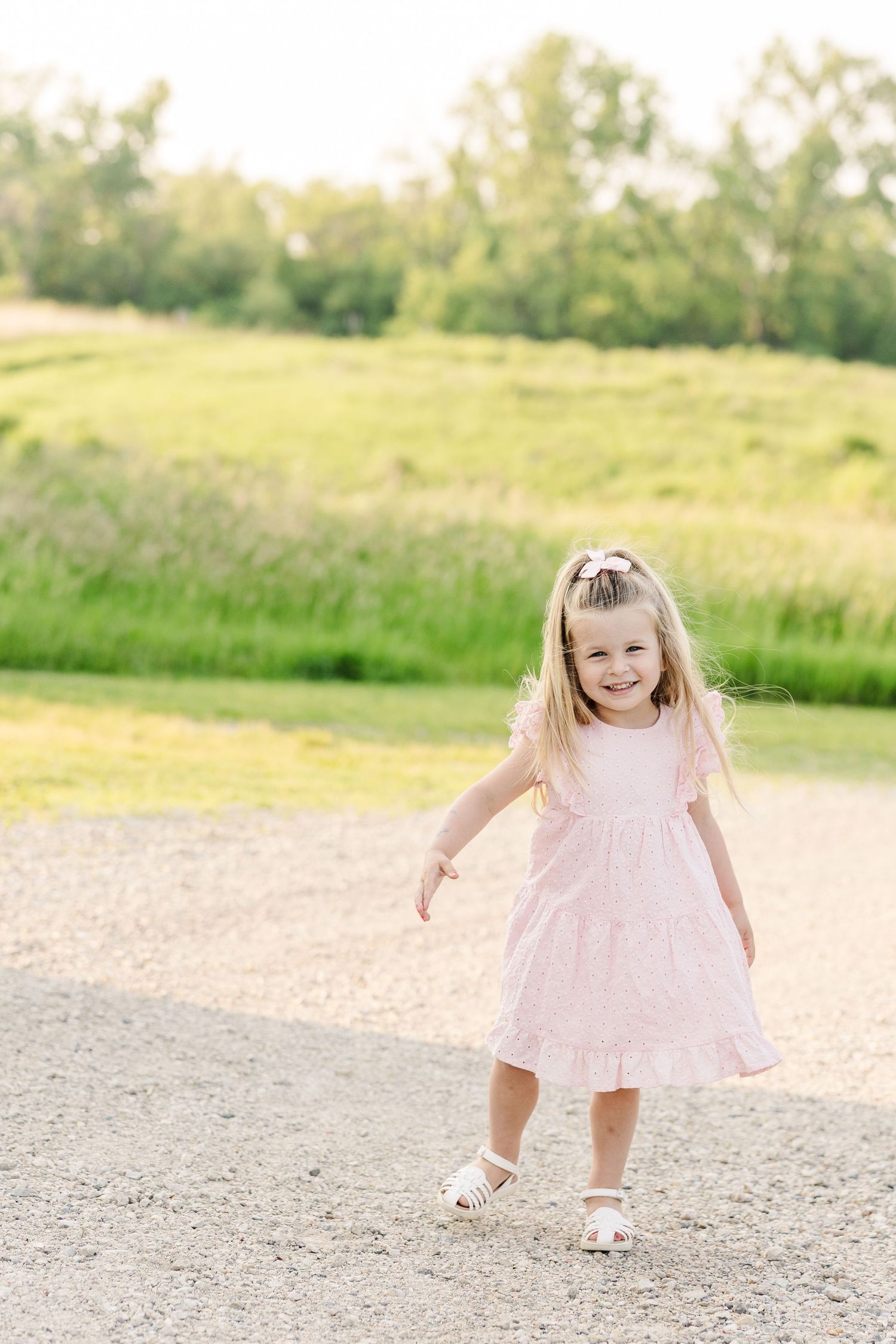 Happy family in coordinating pink and white outfits holds hands with toddler on dirt path during sunset outdoor photo session.