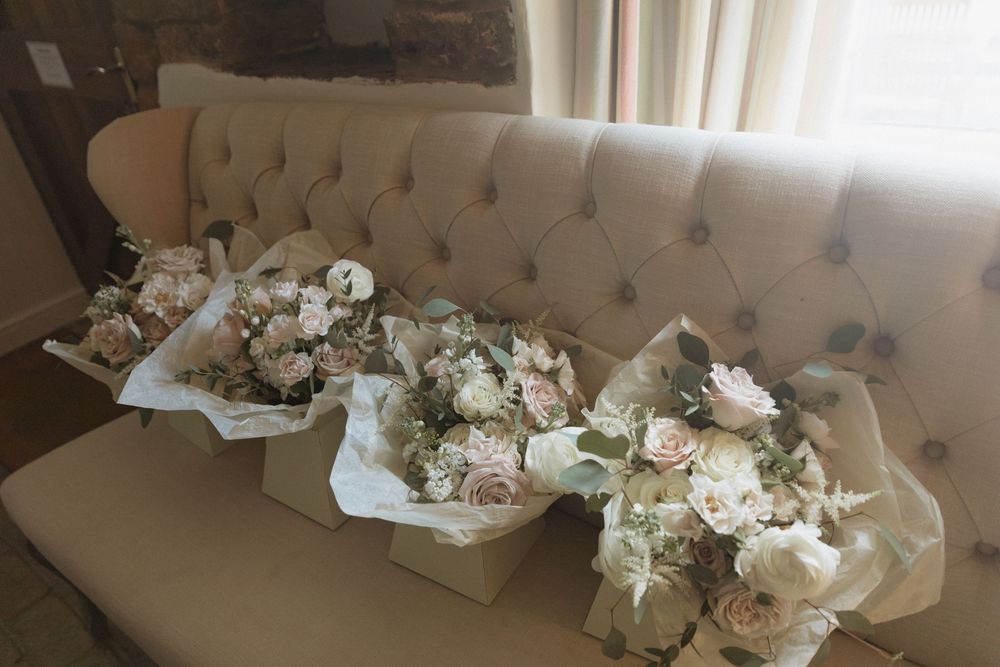 Several elegant white and blush pink wedding bouquets wrapped in tissue paper rest on a tufted beige couch.